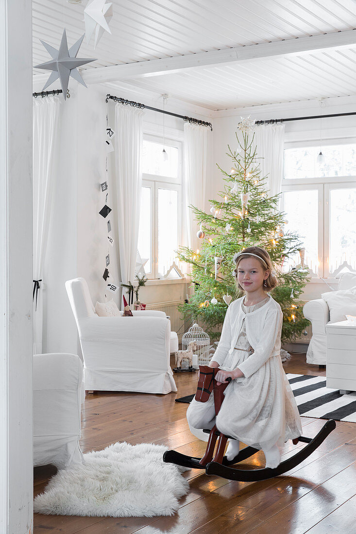 Mädchen mit Schaukelpferd im Wohnzimmer, im Hintergrund Weihnachtsbaum