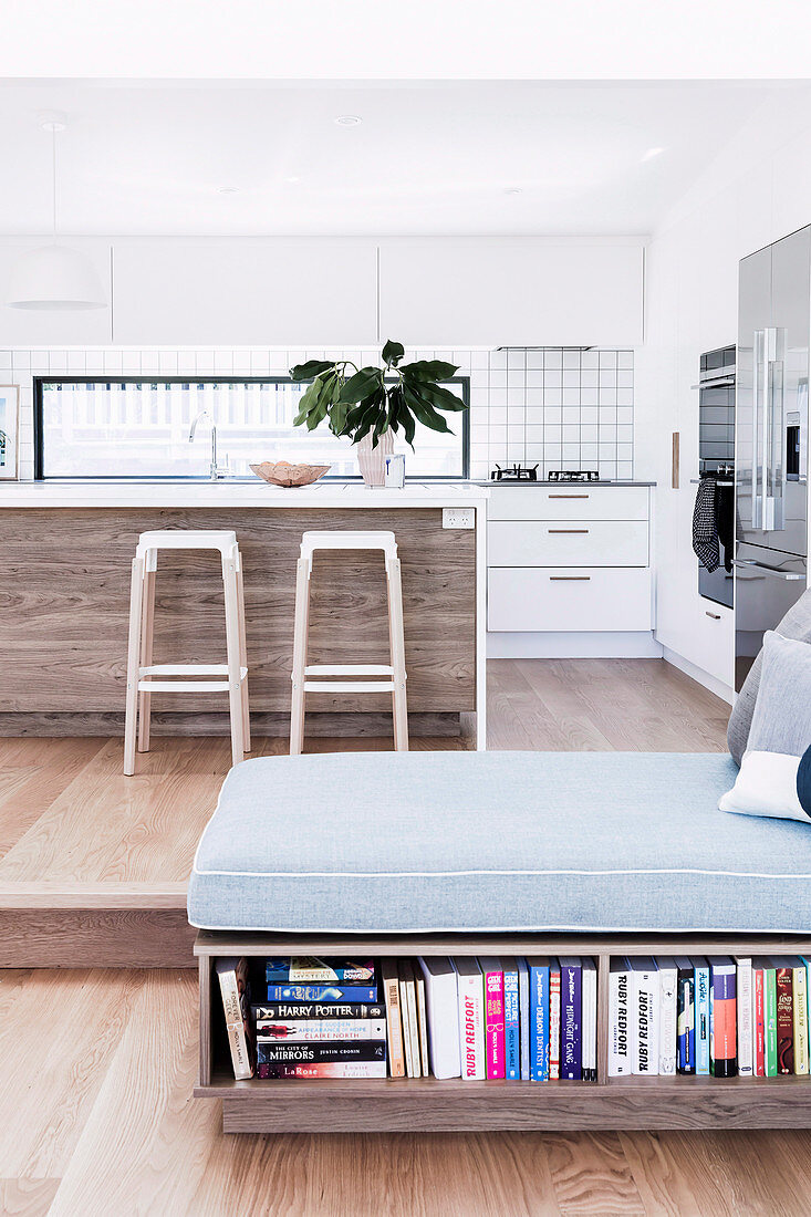 Open kitchen with island and bar stools on pedestal, day bed in the foreground with integrated bookcase