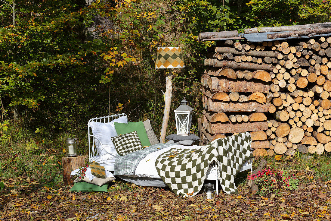 Knitted blanket and cushion on bed and standard lamp in front of stacked firewood on edge of forest