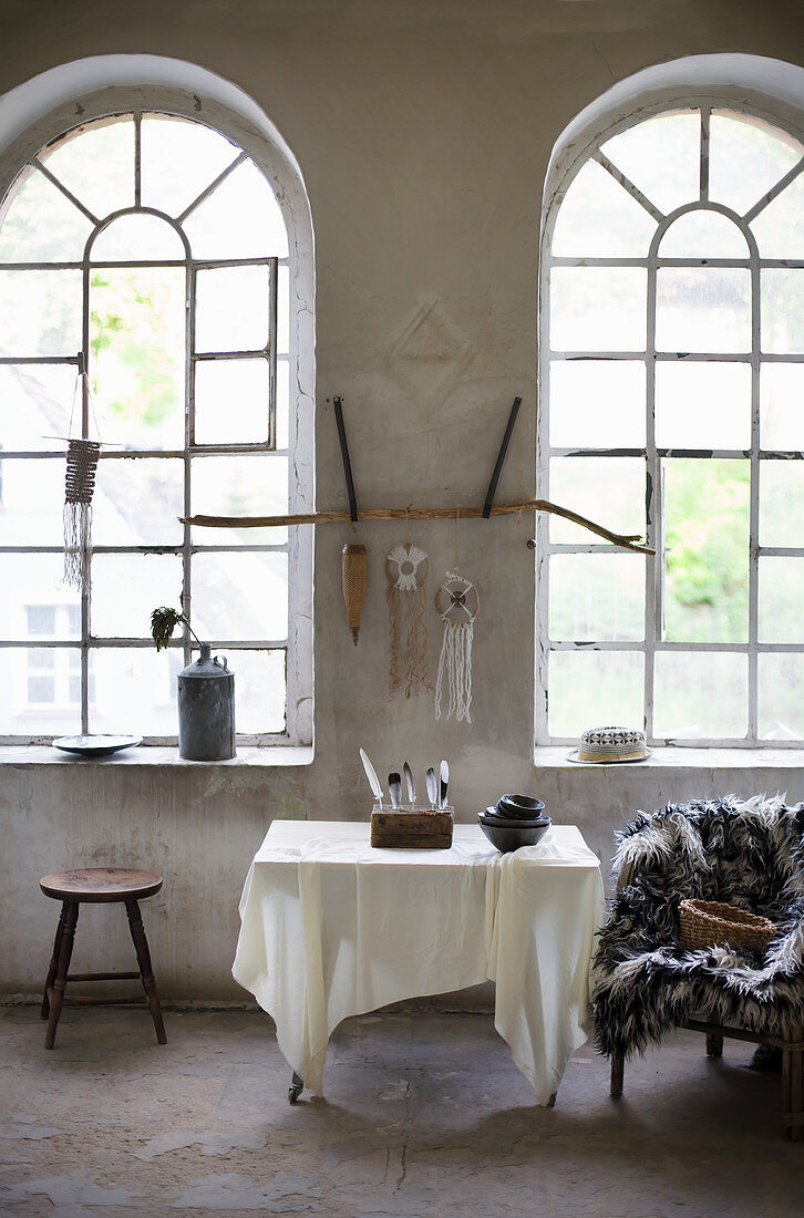 Tisch mit Schalen und Federsammlung, Sessel mit Felldecke und Hocker vor Rundbogenfenster im Loft