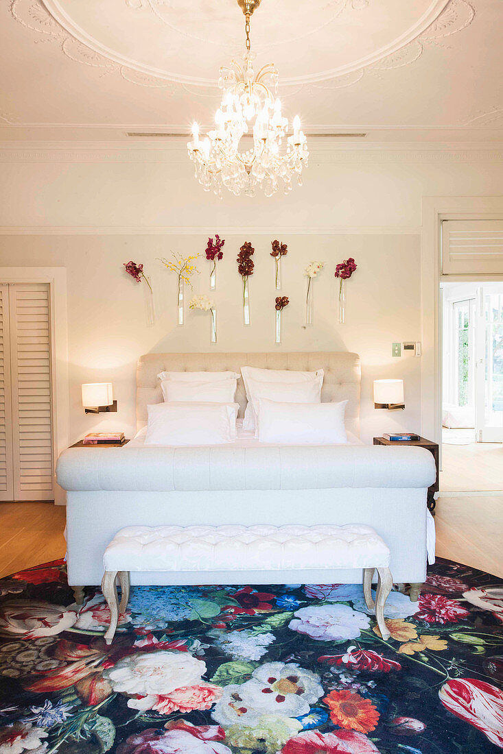 Elegantes Schlafzimmer mit weißem Doppelbett, Kleiderbank, Kronleuchter und Blumen an der Wand