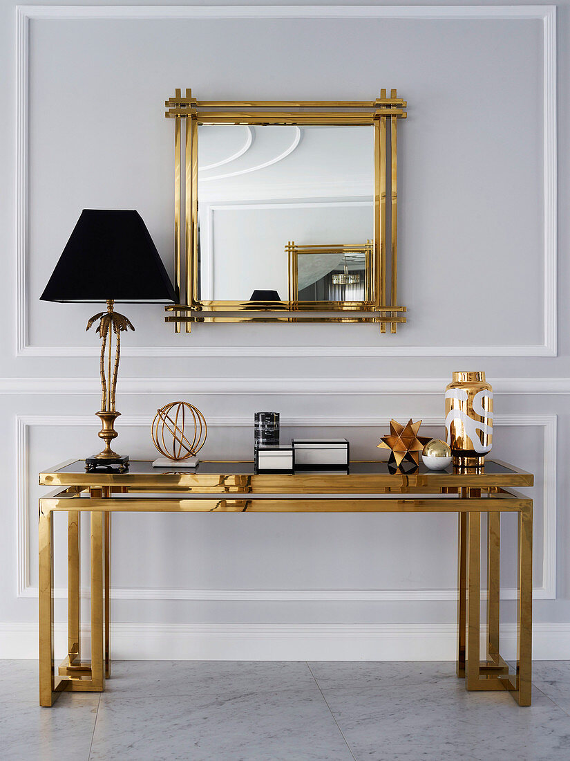 Table lamp on console with gold-colored frame and glass top, above wall mirror with gold frame