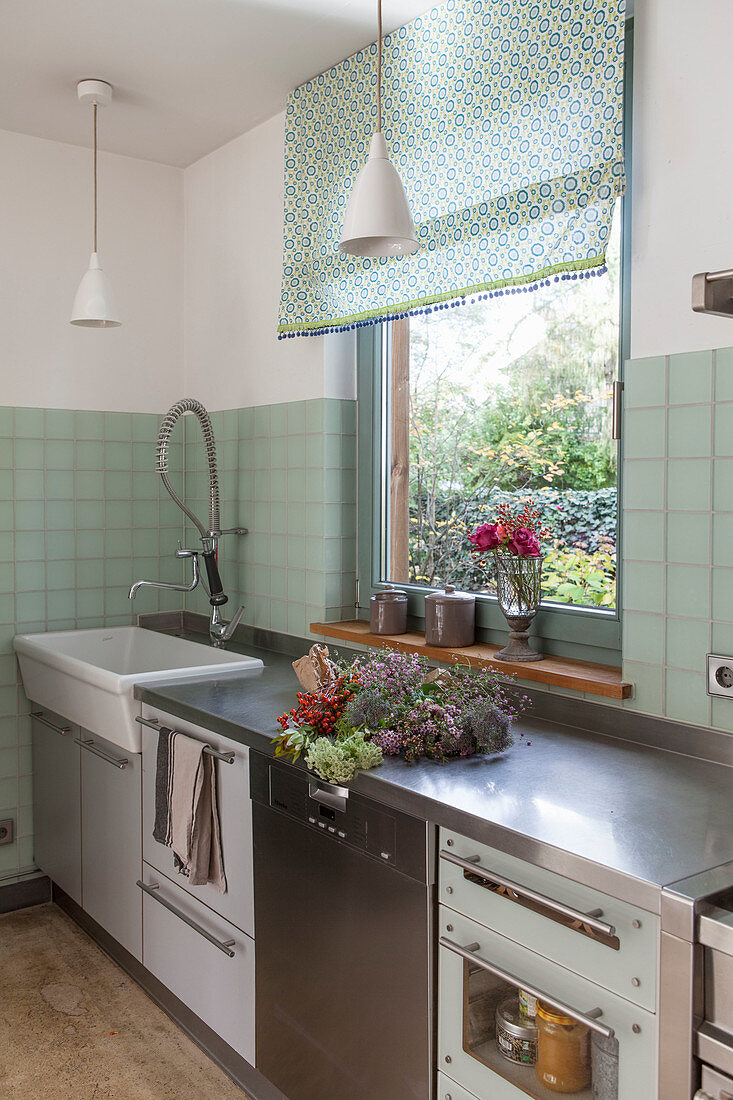 Green wall tiles and kitchen counter with stainless steel worktop below window in kitchen