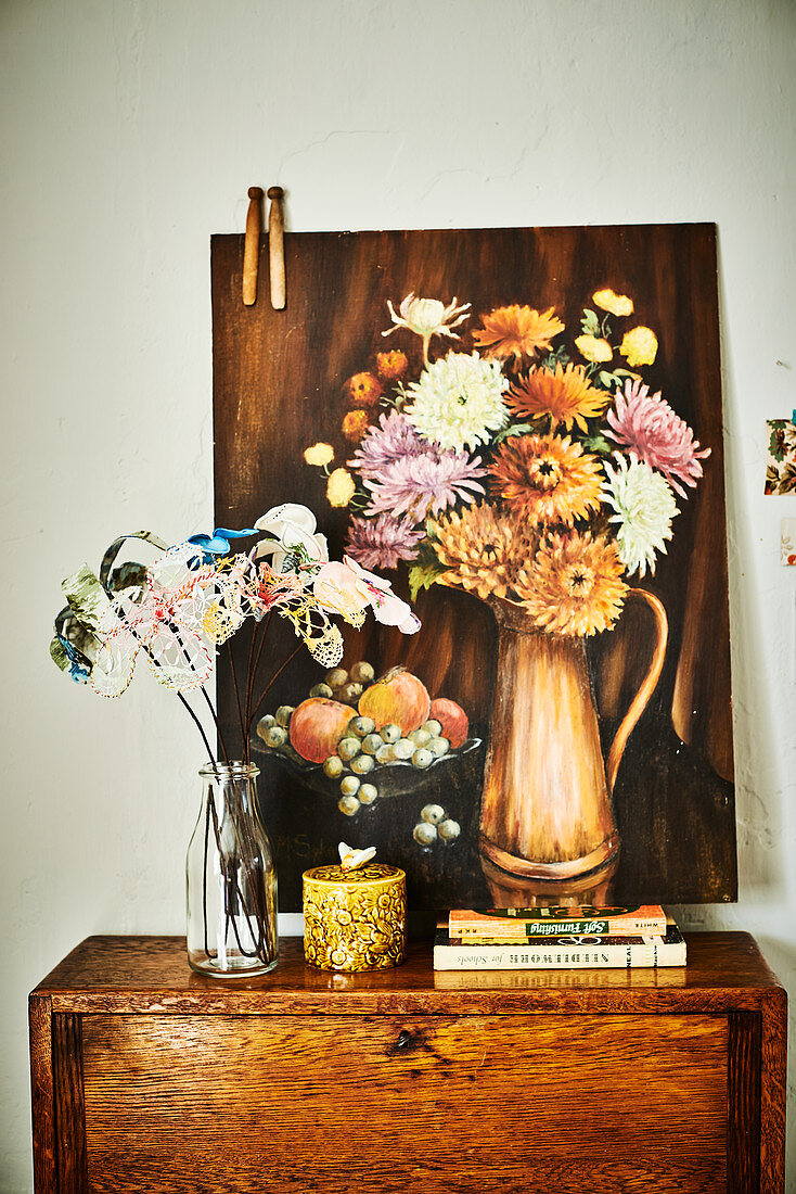 Glasflasche mit Stoffblumen auf Kommode, Bild mit Blumenstillleben an die Wand gelehnt