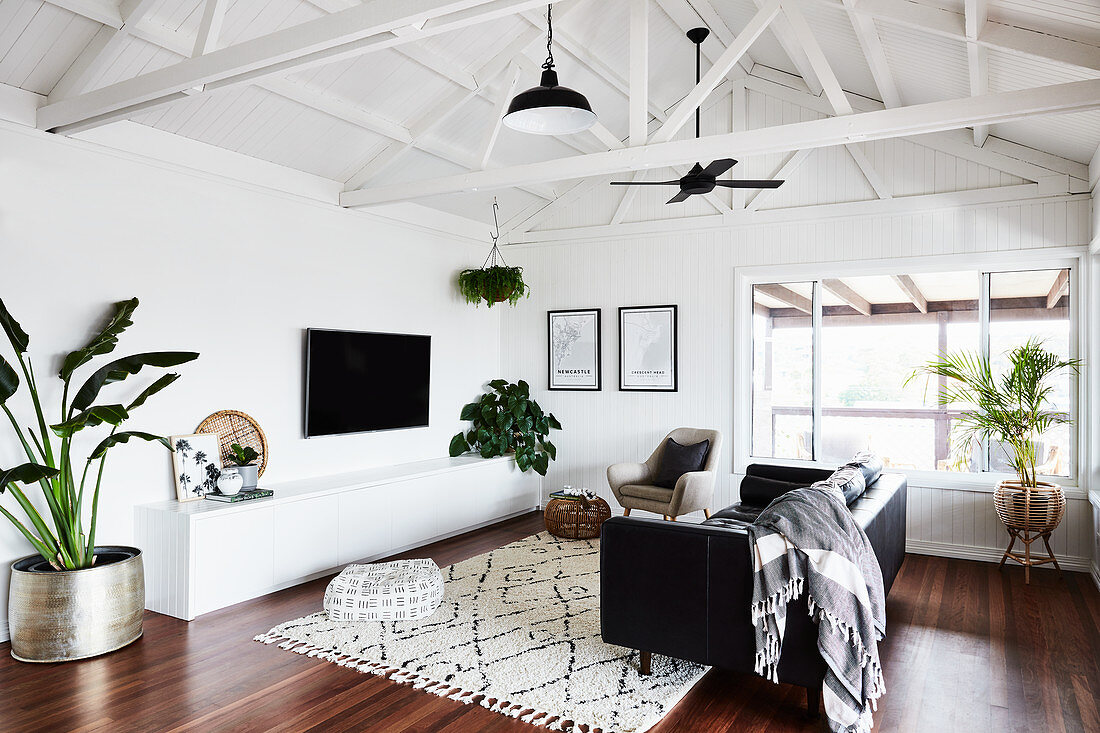 Black leather couch, sideboard, TV and houseplants in living room with white wooden panelling