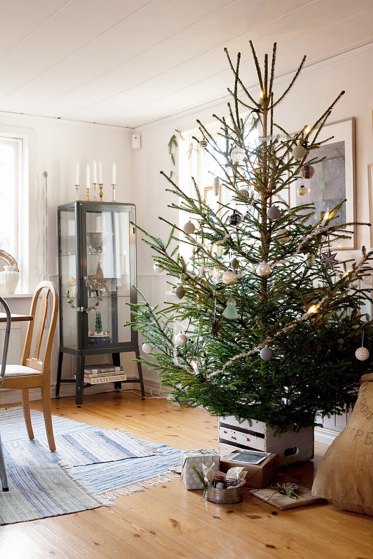 Geschmückter Weihnachtsbaum mit Geschenken, im Hintergrund Vitrinenschrank im Wohnzimmer