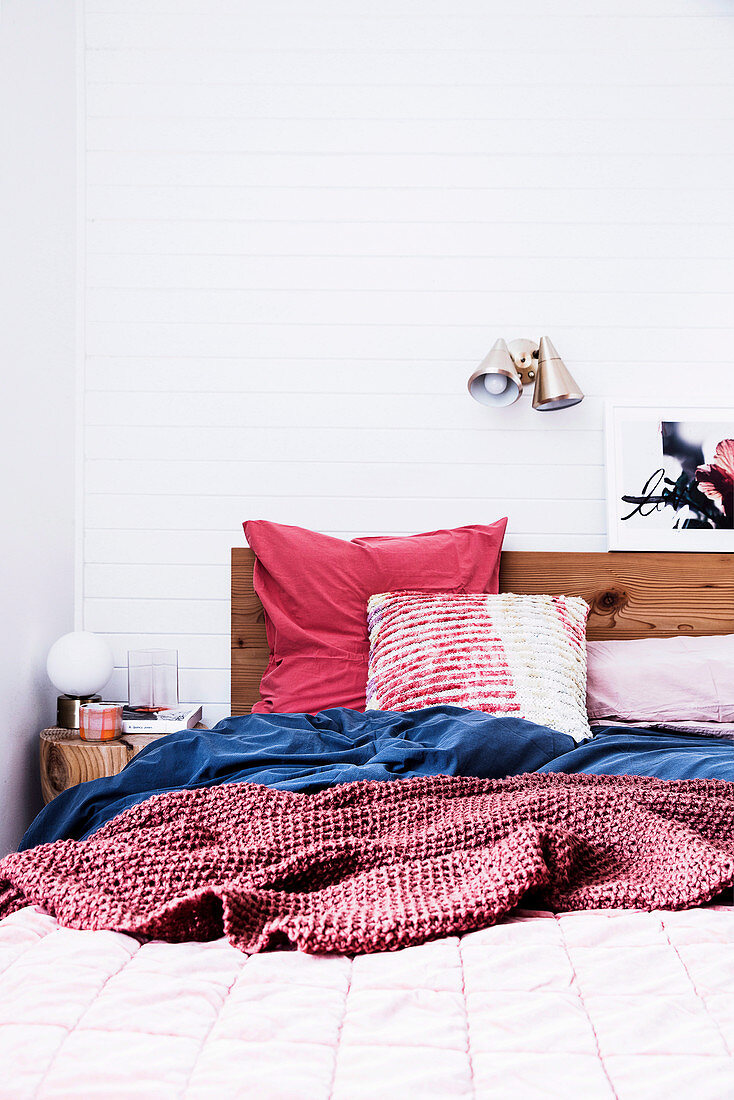Holzbett mit gemütlicher Bettwäsche in Rot und Blau vor weißer Wand