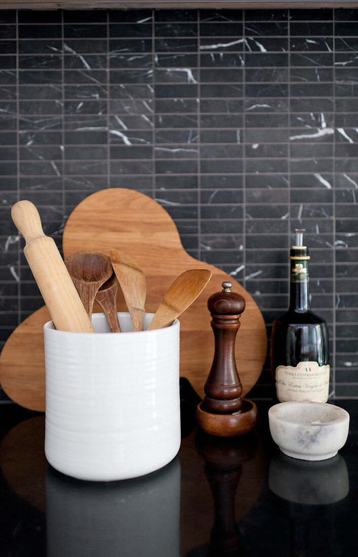 Kitchen utensils, wooden board and pepper mill against black-tiled splashback