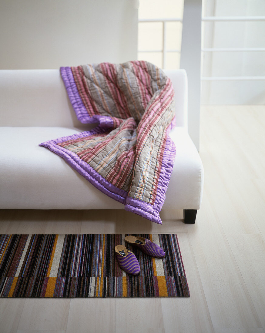 Striped quilted blanket on plain white sofa and purple slippers on striped rug
