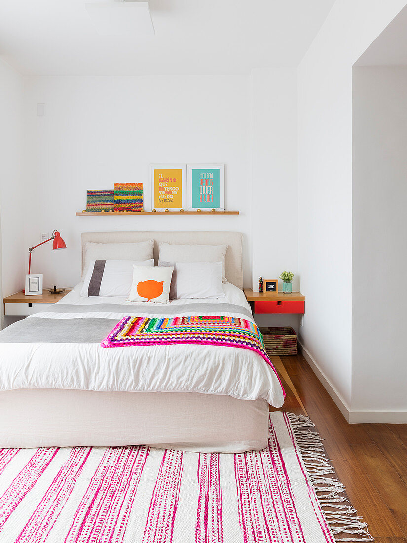 Colourful crocheted blanket on double bed and pink-and-white striped rug in white bedroom