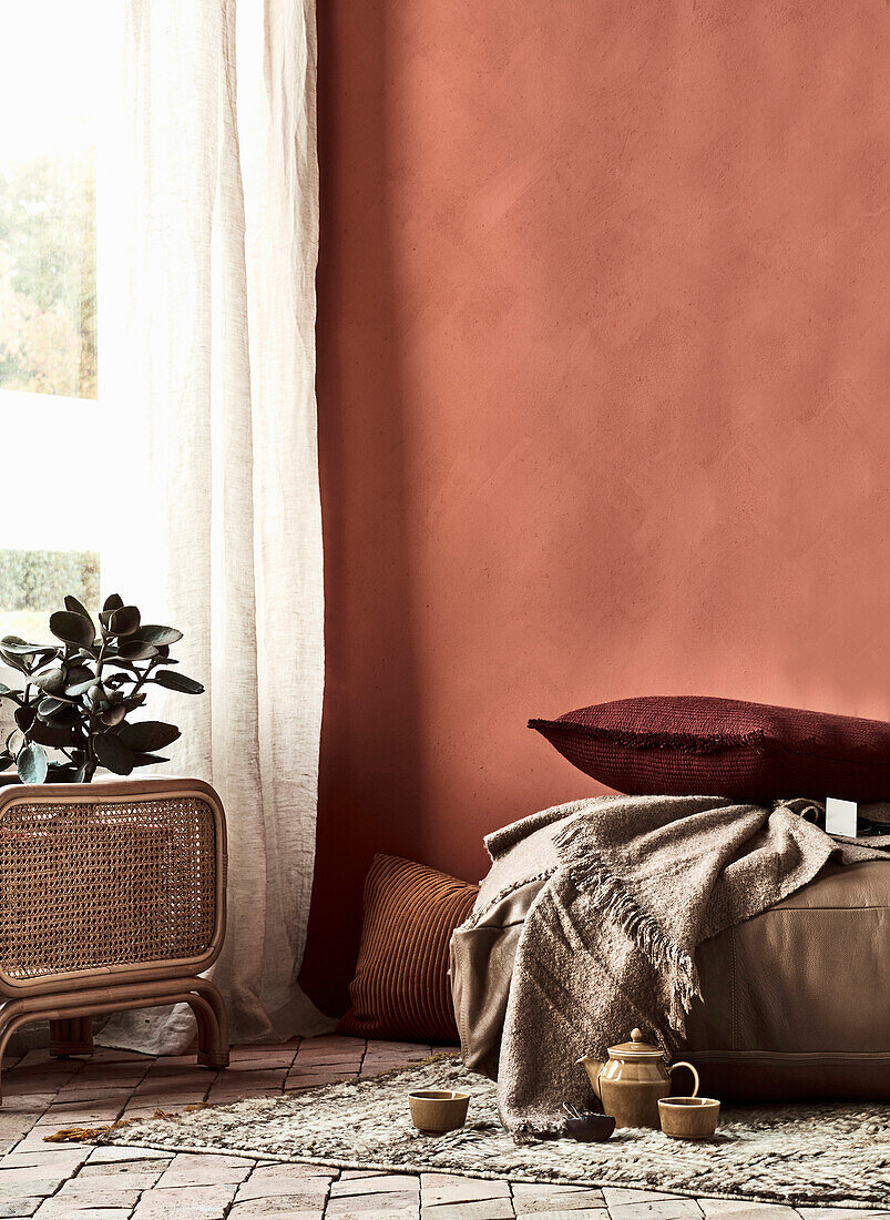 Gemütlicher Sitzkissen und Teegeschirr auf Teppich vor terrakottafarbener Wand, Zimmerpflanze am Fenster