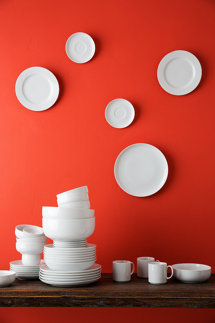 Weisses Geschirr auf Tisch und weiße Teller an roter Wand dahinter