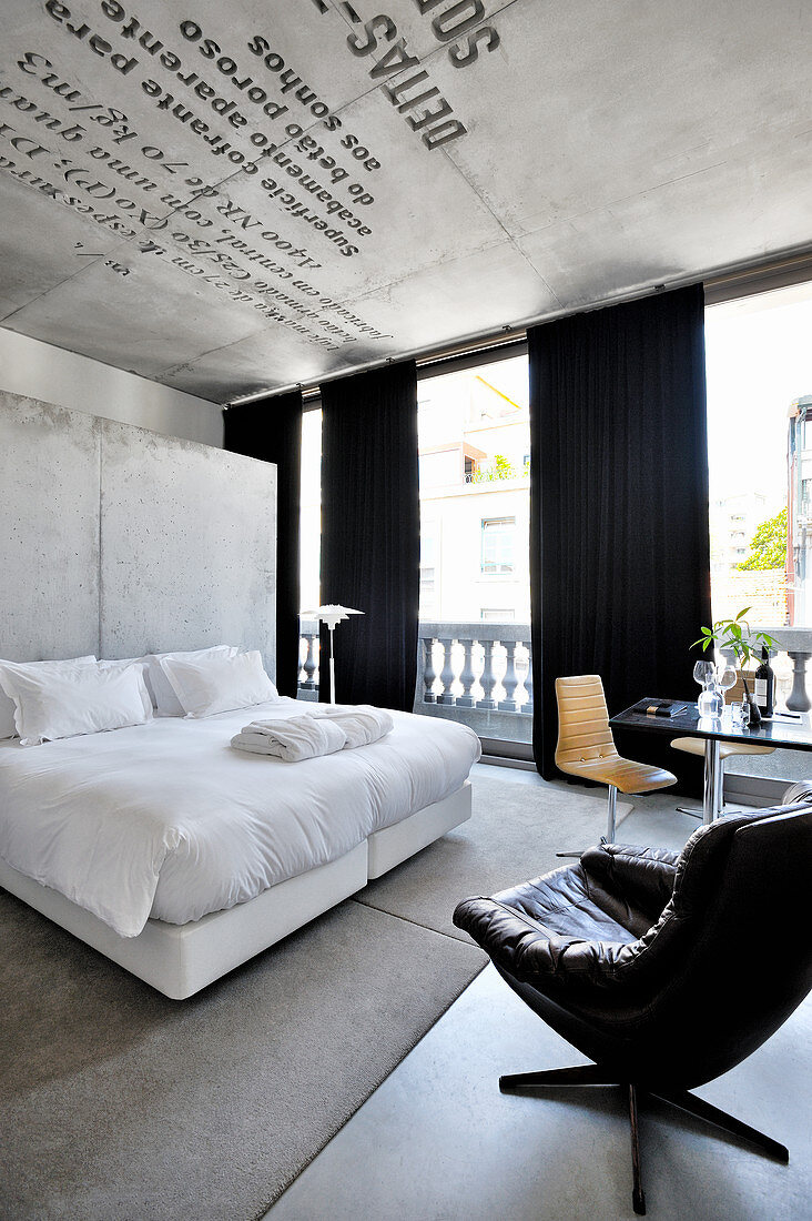 Doppelbett mit weißer Bettwäsche vor Betonwand im Hotelzimmer, im Vordergrund Ledersessel
