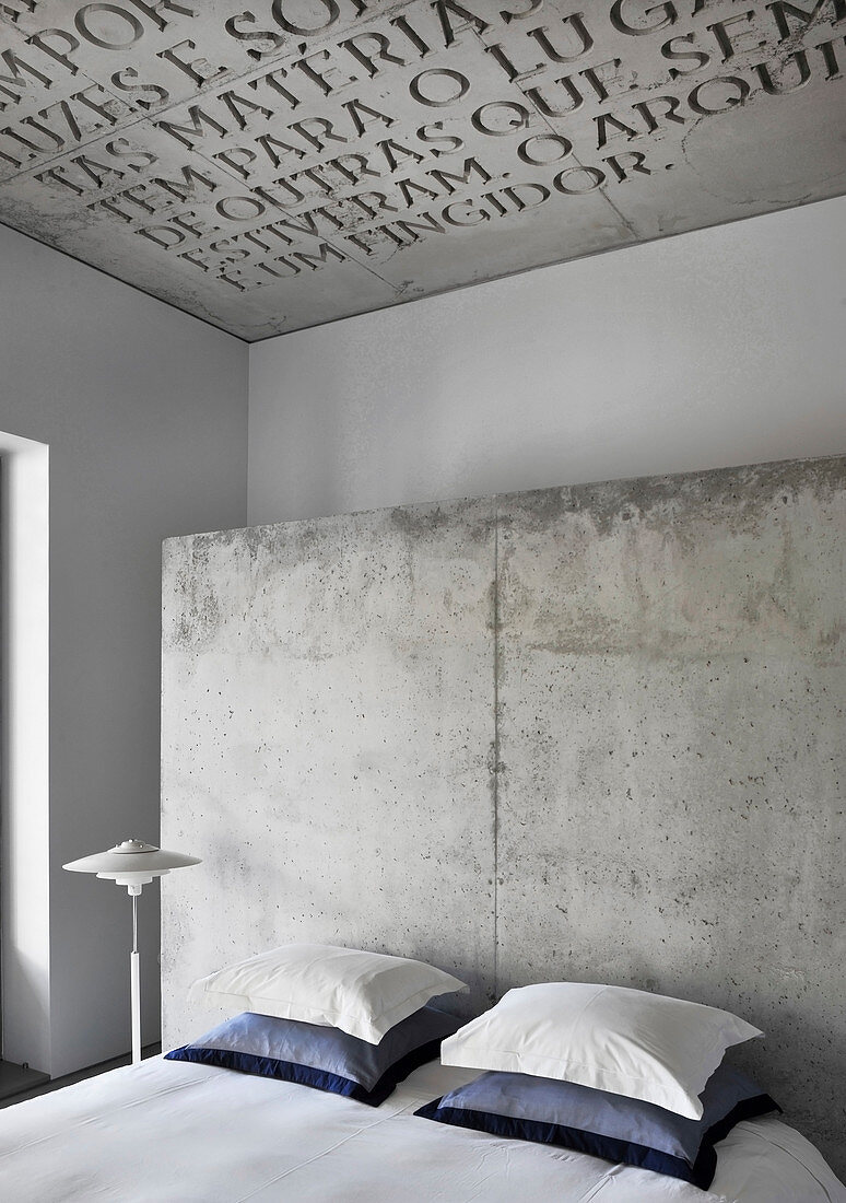 Double bed with pillows against concrete partition in hotel room with lettering on concrete ceiling