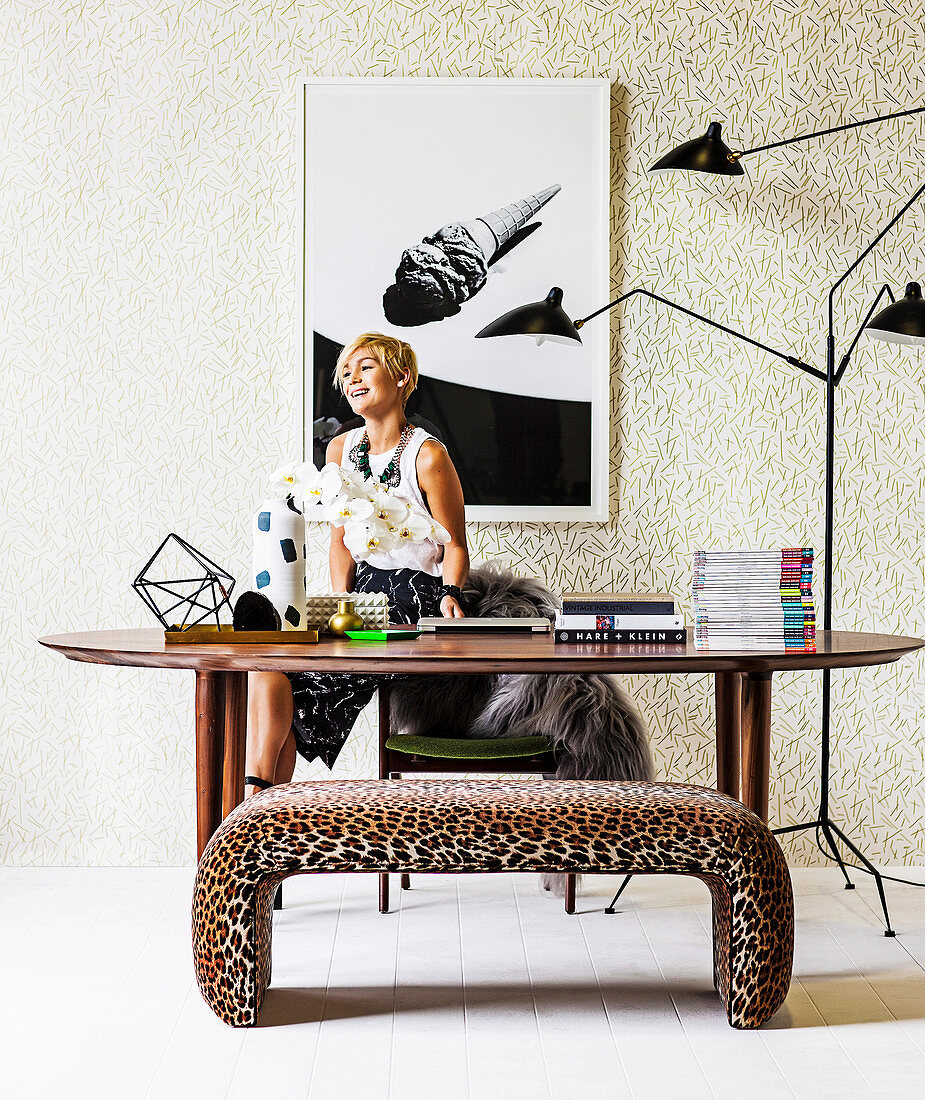 Blonde Frau hinter ovalem Esstisch, mehrarmige Stehleuchte und Sitzbank im Leoparden-Look