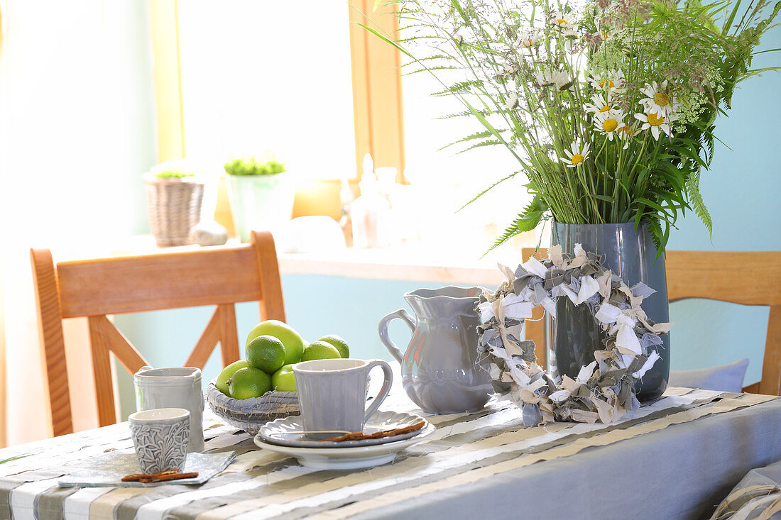 DIY-Tischdecke und Kranz mit Stoffbändern, Geschirr und Wiesenblumenstrauß auf Tisch