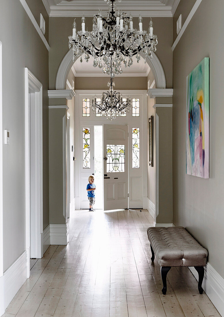 Upholstered bench and chandelier in the hallway with wooden floorboards and arches, toddler in the background
