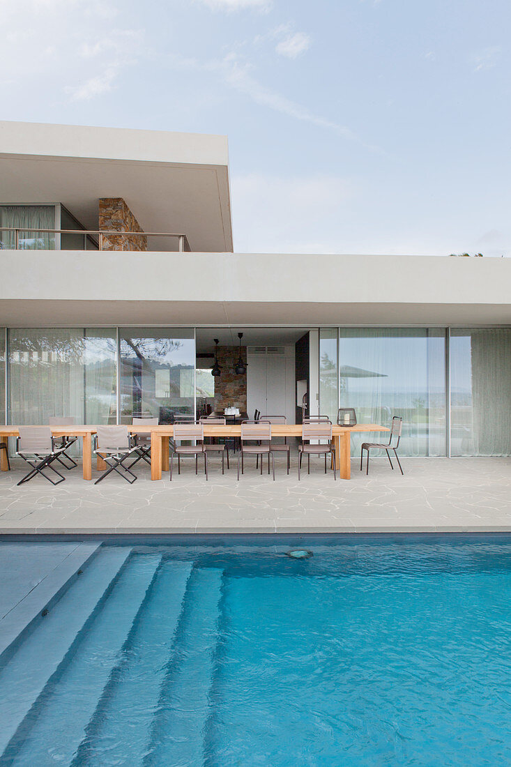 Blick über Pool auf Terrasse mit Natursteinplatten, langer Holztisch vor Glasschiebetür