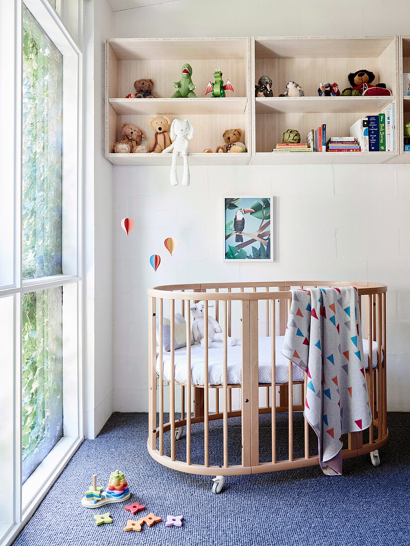 Babybett und Regal vor raumhohem Fenster im Kinderzimmer, Spielzeug auf dem Teppich