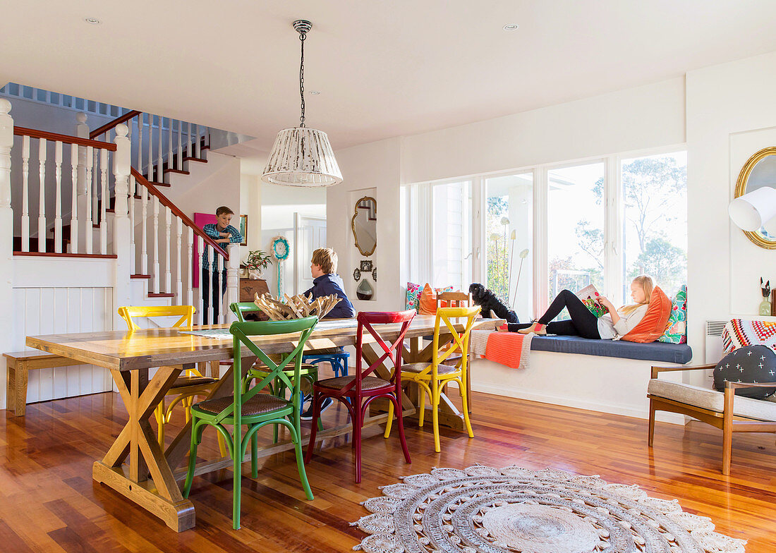 Long dining table with colorful chairs in an open living room with stairs, family in the room