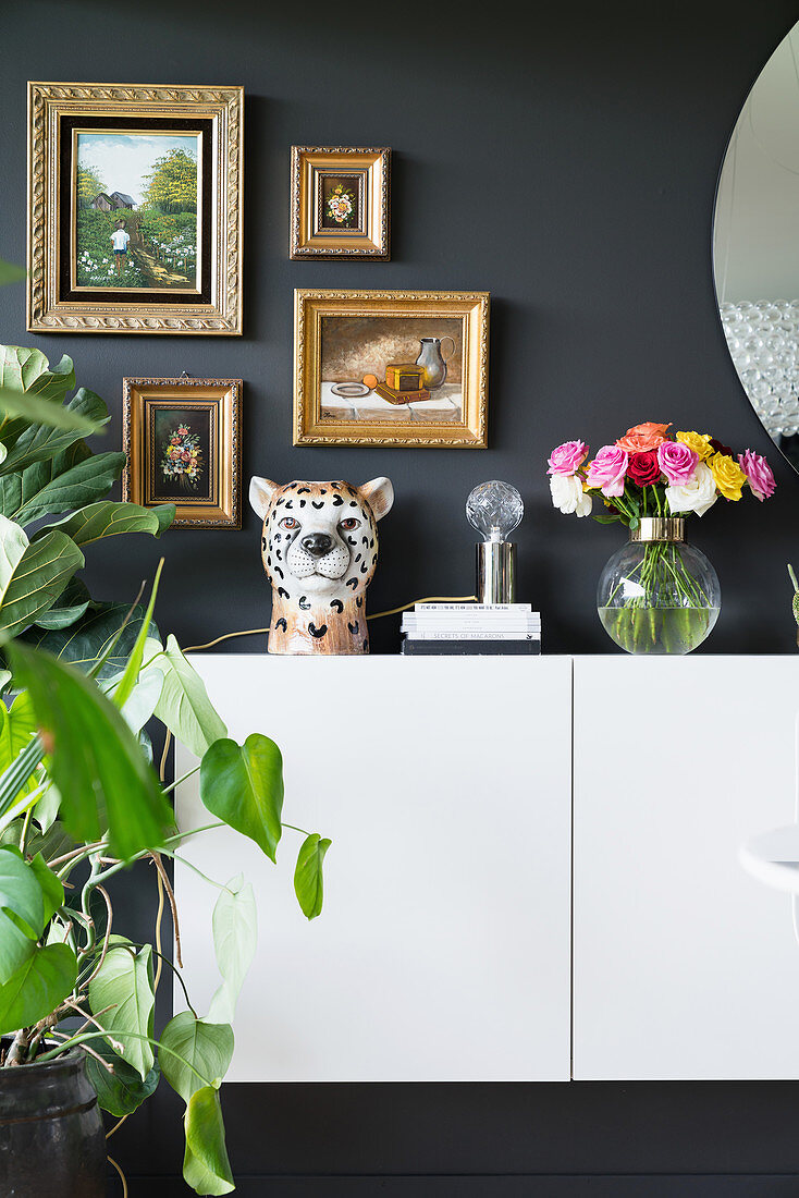 Leopardenkopf aus Keramik und Blumenstrauss auf weissem Sideboard, darüber Bilder in Goldrahmen an dunkler Wand
