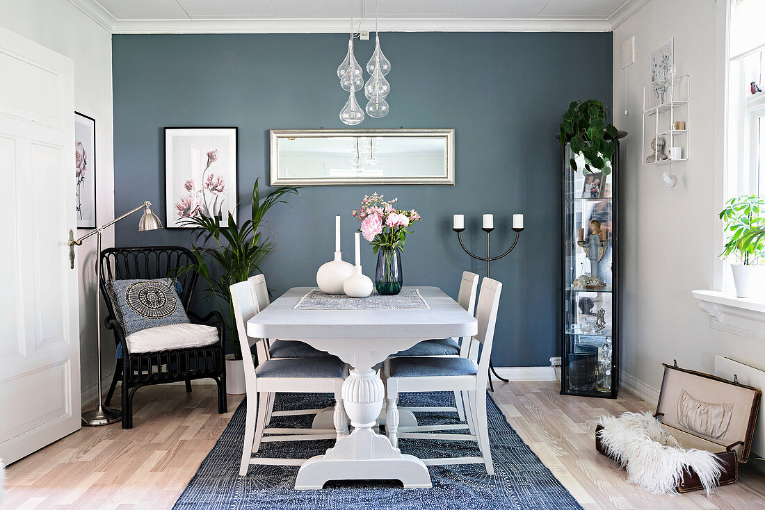 Weißer Tisch mit Stühlen, Vitrinenschrank und schwarzer Korbstuhl im Essbereich mit matt-blauer Wandfarbe