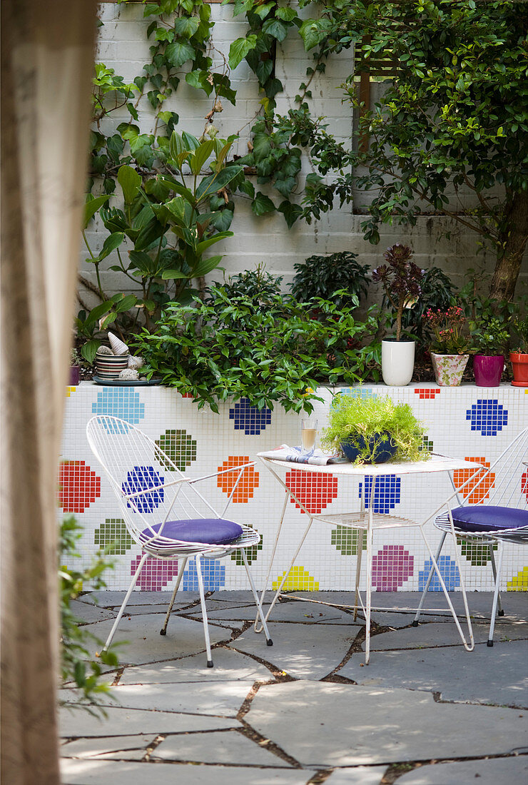 Delicate metal table and two chairs on terrace with multi-coloured mosaic wall