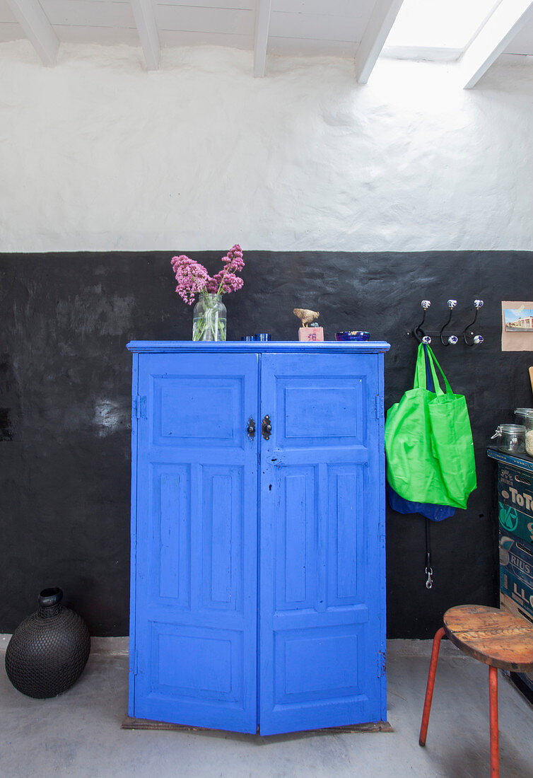 Blauer, halbhoher Schrank, daneben Einkaufstaschen auf Wandhaken und schwarze Bodenvase vor schwarzer Wand