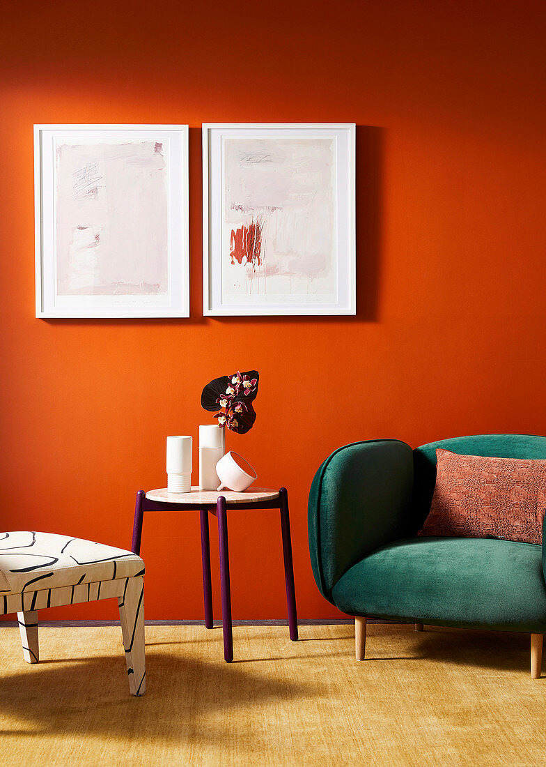 Stuhl, runder Tisch und Sessel mit grünem Samtbezug vor orangefarbener Wand mit Bildern