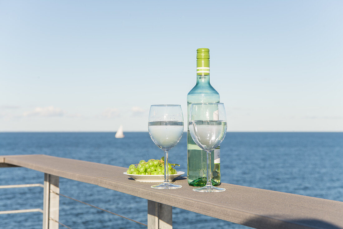 Weinflasche mit Gläsern und Trauben auf Terrassenbrüstung mit Blick aufs Meer