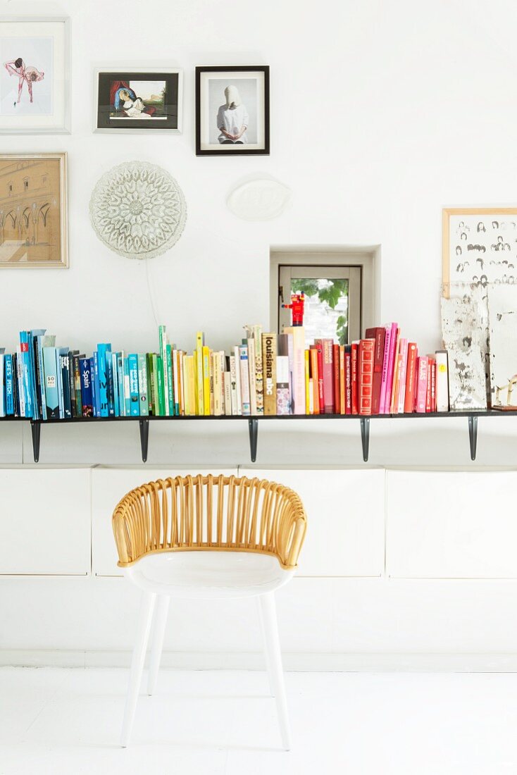 White wicker chair in front of books arranged by colour on wall-mounted shelf