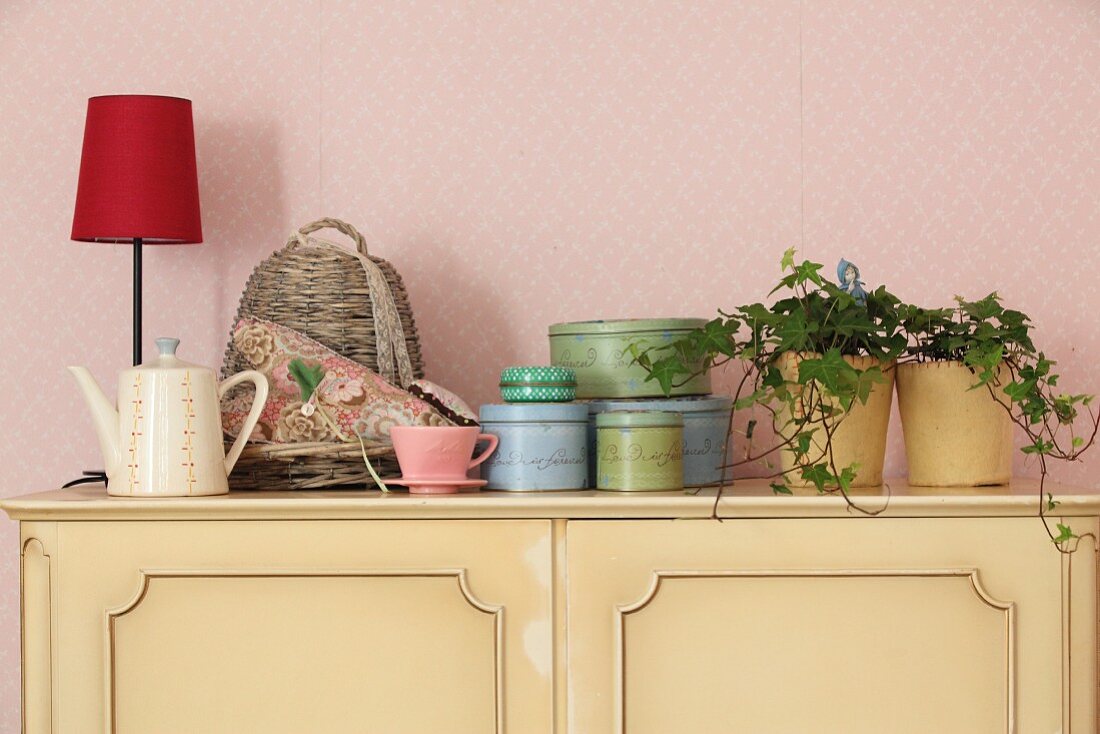 Zimmerpflanzen, Blechdosen und Tischlampe auf pastellgelber Kommode