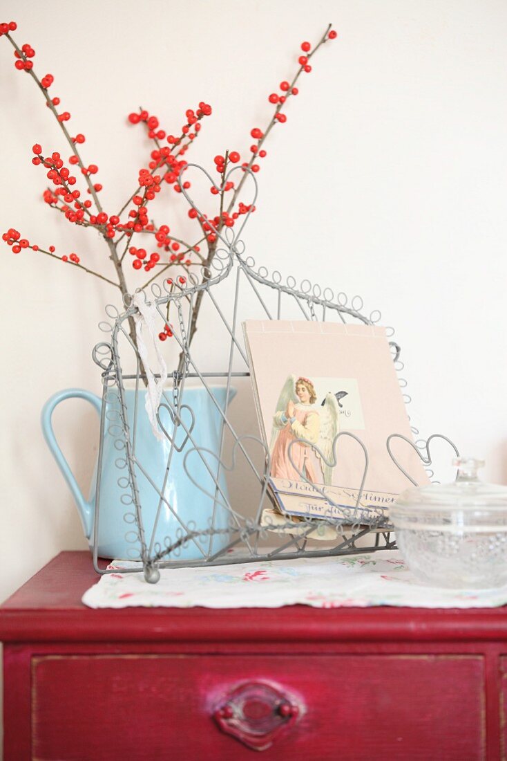 Branches of red berries in pale blue jug and vintage angel motif on wire rack on top of vintage chest of drawers