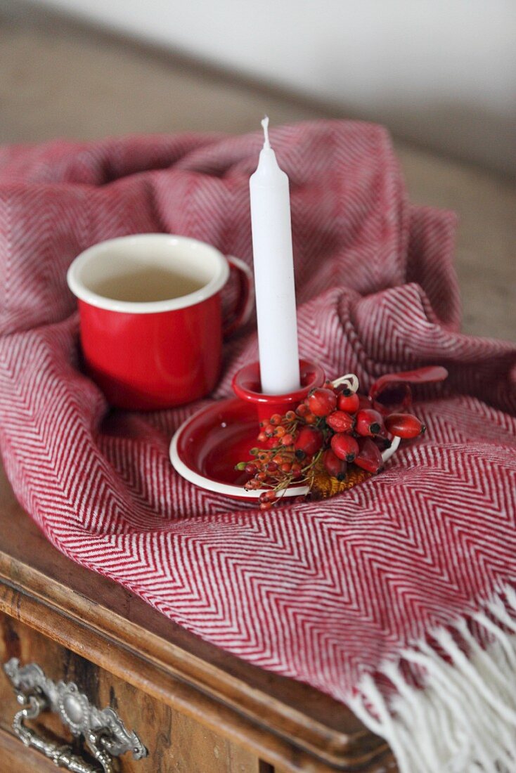 White candle in red candlestick with rose hips next to mug on fringed blanket