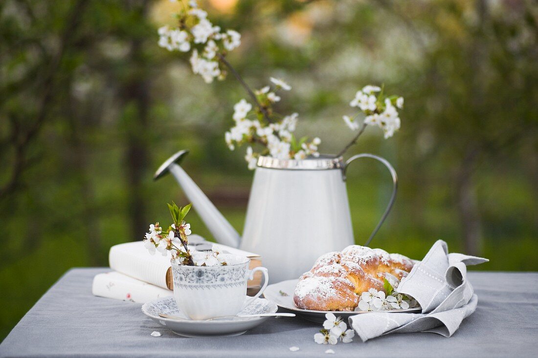 Kaffeetasse, Bücher, Hefezopf und Giesskanne mit Blütenzweig auf grauem Tischtuch