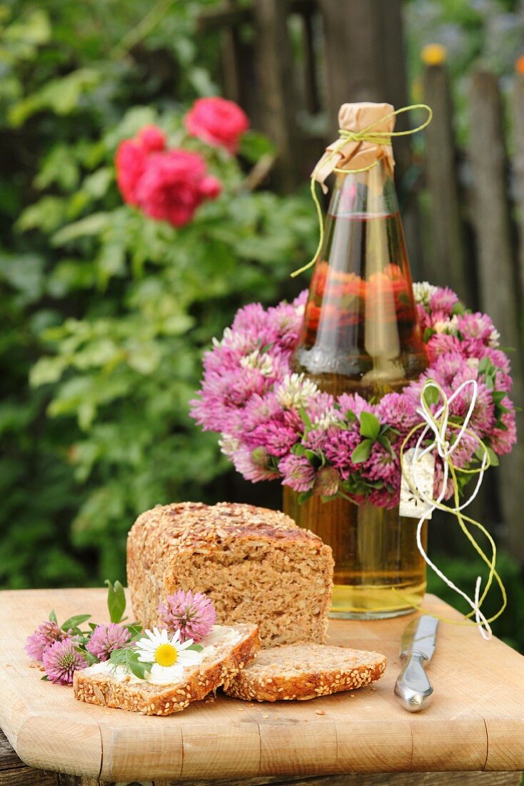 Flasche mit Kleeblütenkranz dekoriert und mit Brot und Essblüten auf Holzbrett