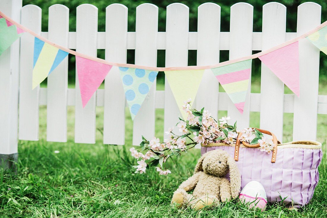 Cloth bunny and Easter eggs below pastel bunting hung on white picket fence in garden