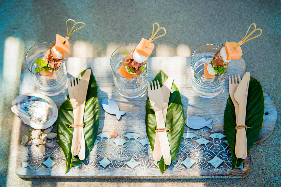 Wooden cutlery on leaves and summery skewers in glasses on tray
