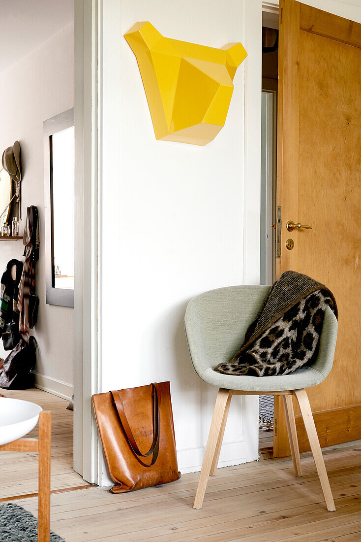 Stylised yellow bear's head on wall above shell chair and leather bag