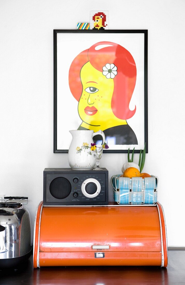 Colourful artwork above radio and fruit basket on top of old bread bin