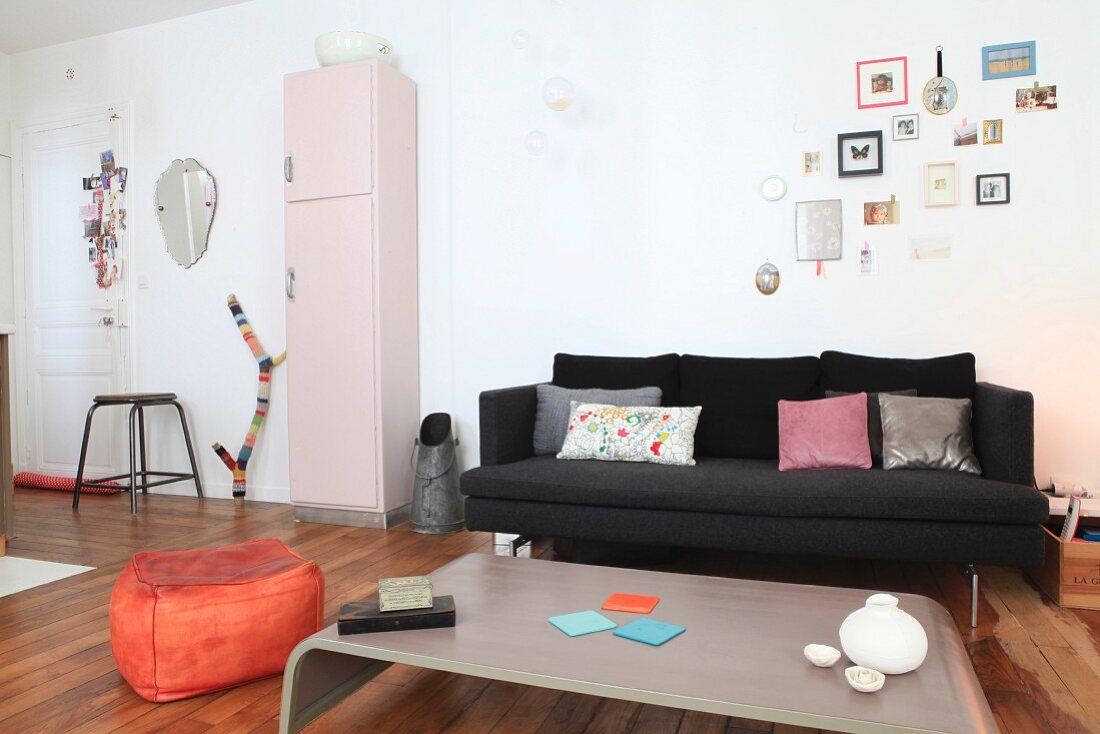 Offener Wohnbereich mit schwarzer Couch, Kohlenschütte, rosafarbenem Retroschrank und orangefarbenem Sitzpouf