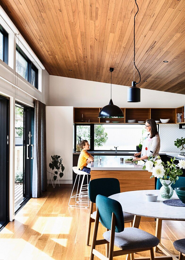 Offene Küche und Essbereich mit Parkettboden, Holzdecke und Terrassentür, im Hintergrund Mutter und Sohn
