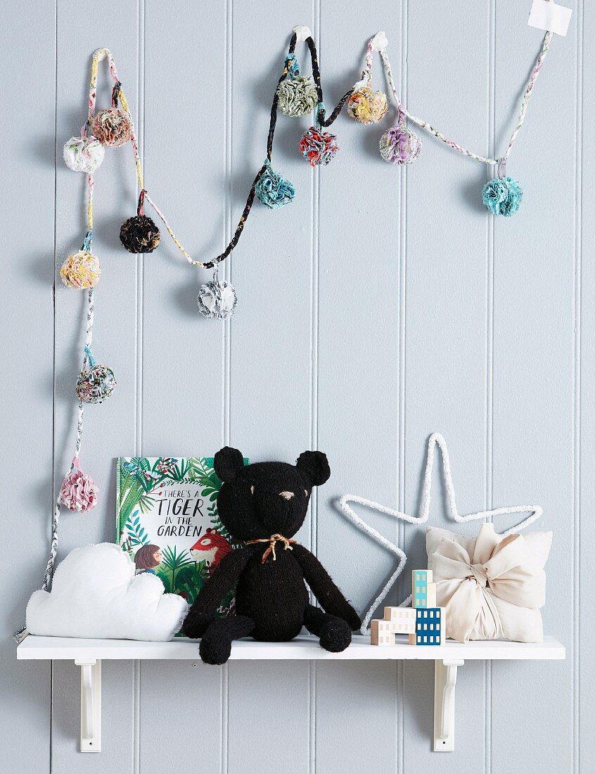 Black, knitted cuddly toy on a white wall shelf under a colorful pompom chain on wooden paneling