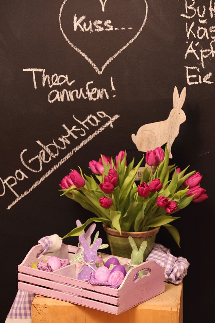 Vase of tulips and Easter ornaments in wooden tray against chalkboard wall with messages in chalk