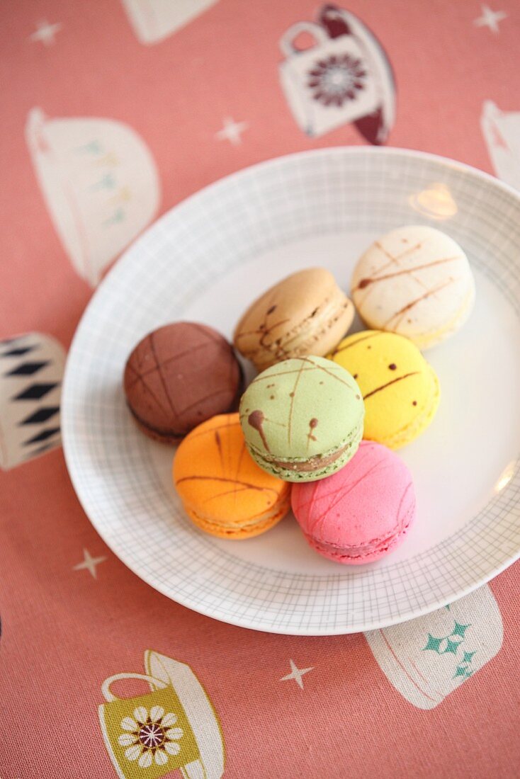 Teller mit bunten Macarons auf Tischdecke mit Kaffeetasse-Motiven