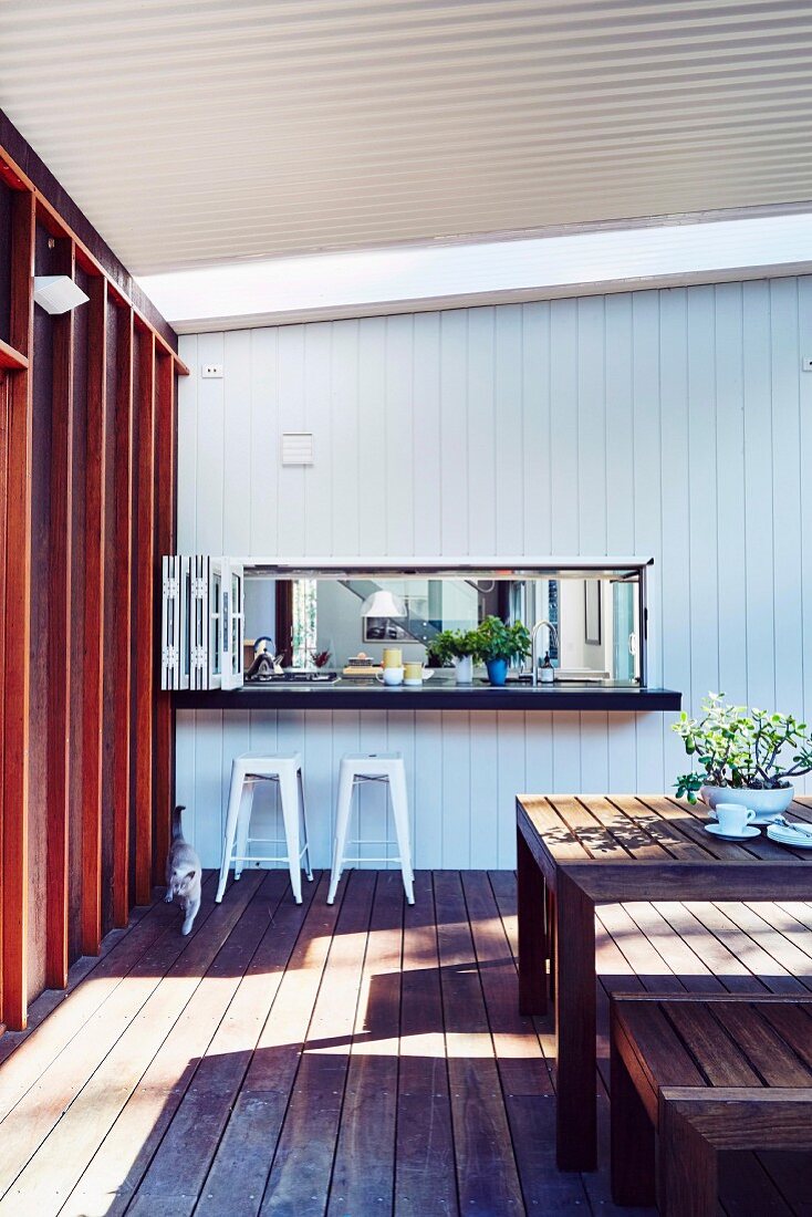 Tisch mit Bank und Barhocker vor Durchreiche auf überdachter Terrasse mit Holzverkleidung