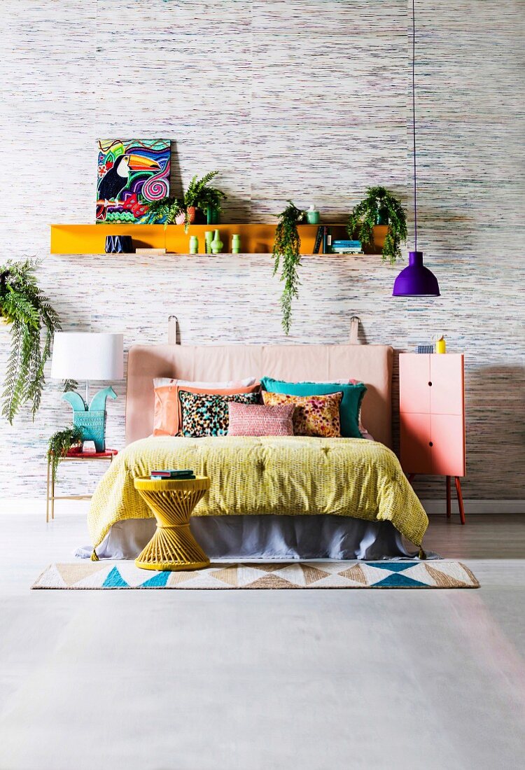 Colorful blanket and pillows on bed with leather headboard, pendant lamp with purple lampshade and shelf with picture and house plants against wallpaper