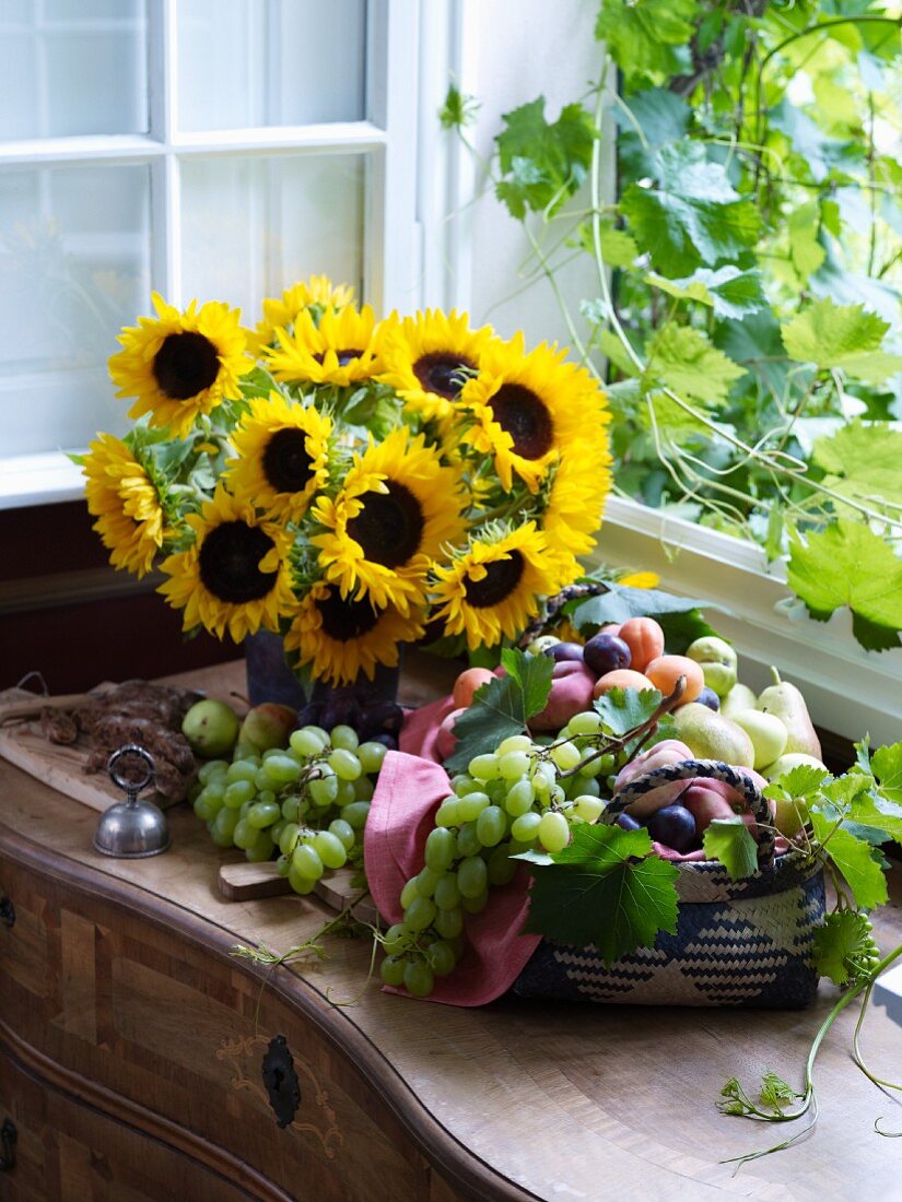 Opulent fruit bowl and vase of sunflowers next to window