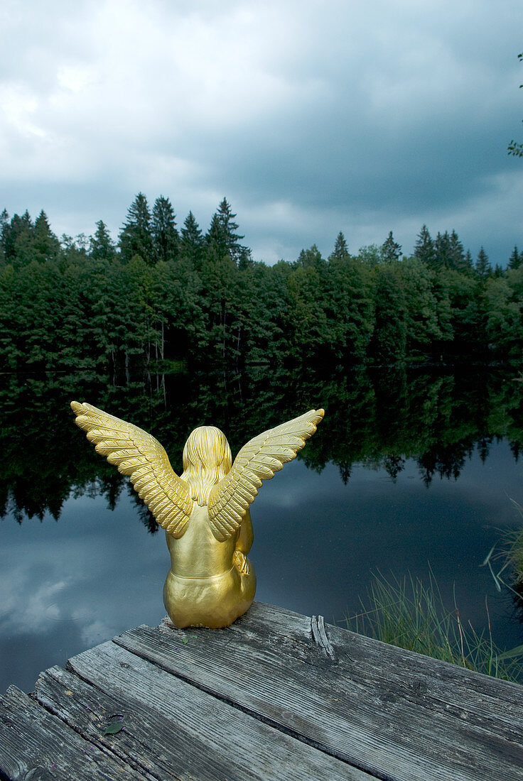 Goldene Engelsfigur sitzt auf dem Steg an einem See im Wald