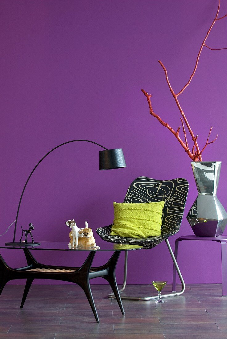 Glastisch mit Bogenlampe und bedruckter Sessel, Keramikvase mit Silberglasur auf Beistelltisch vor lila Wand